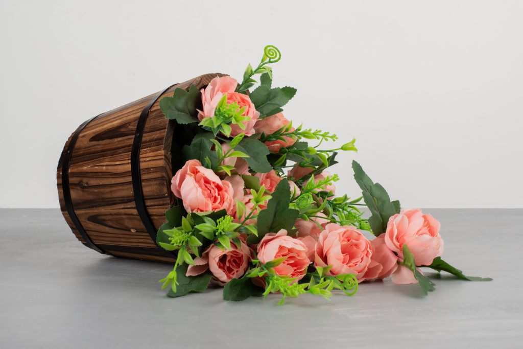 Realistic fake pink roses spilling out of a rustic wooden bucket on a gray surface, best fake wedding flowers.