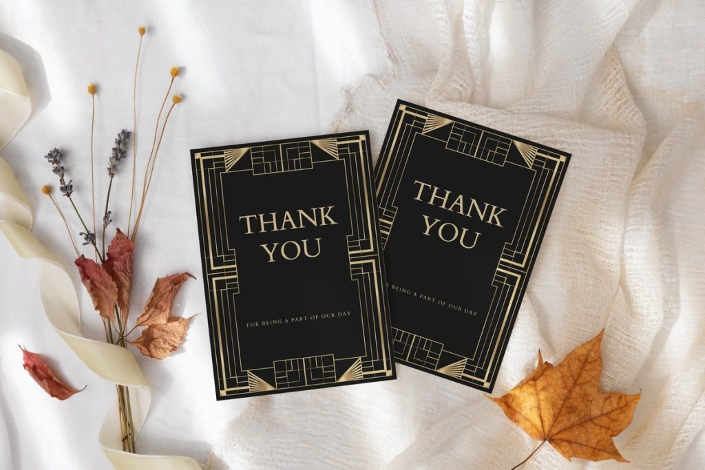 Two black-and-gold art deco wedding thank you cards with geometric borders on soft white fabric, styled with dried autumn leaves and florals.