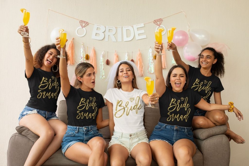Bride bachelorette party shirts with the bride and bridesmaids celebrating together on a sofa.