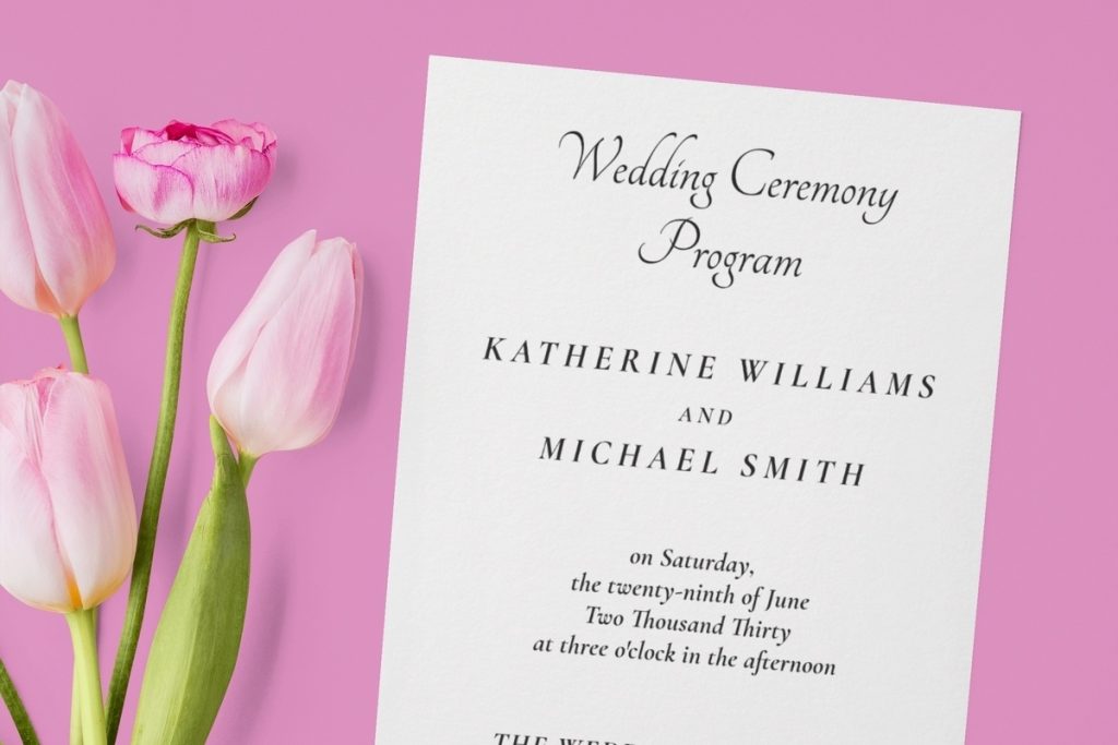 Custom wedding programs with elegant black script text, displayed on white cardstock next to pink tulips on a pastel pink background.