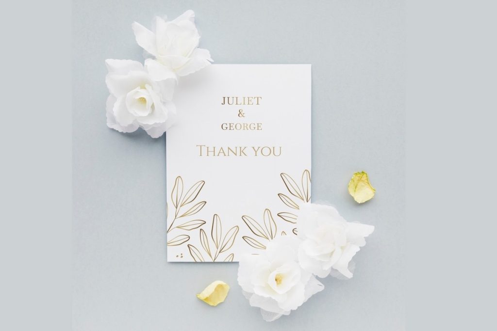 Gold foil wedding thank you cards with botanical design on white background, styled with white flowers and gold petals for an elegant flat lay.