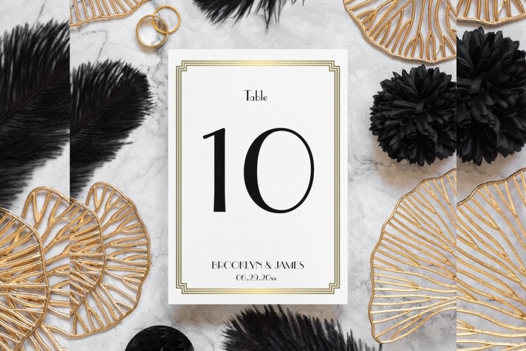Gold Great Gatsby table numbers card with black font, styled among black feathers and gold Art Deco fans.