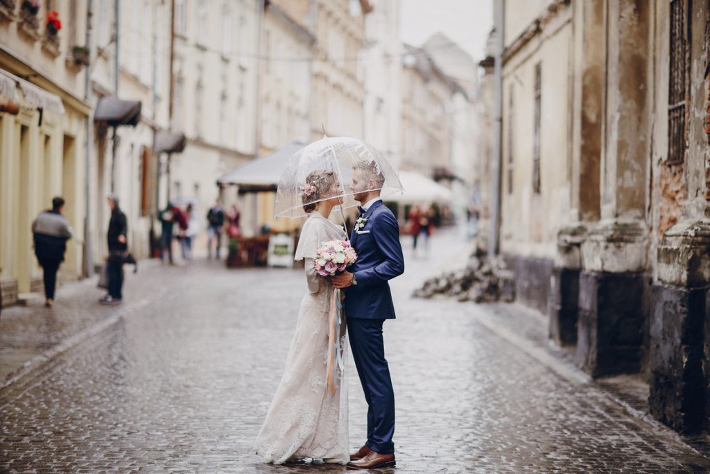Romantic wedding ideas, bride and groom sharing a kiss under an umbrella on a charming cobblestone street.