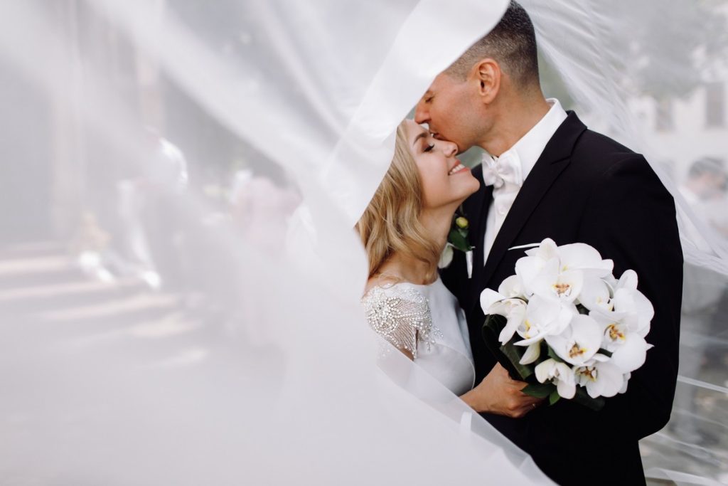 Bride and groom kissing under a flowing veil, groom holding a bouquet of white orchids, About Us Dream Wedding Ideas.