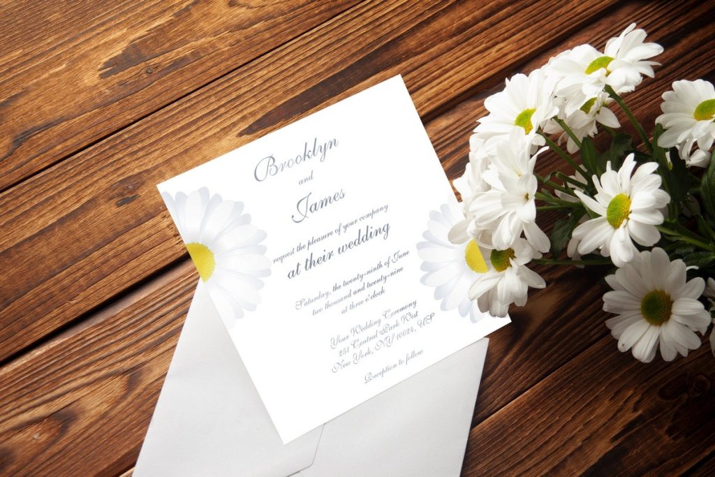 Elegant square daisy themed wedding invitations with white floral design, displayed on rustic wooden table beside fresh daisies.