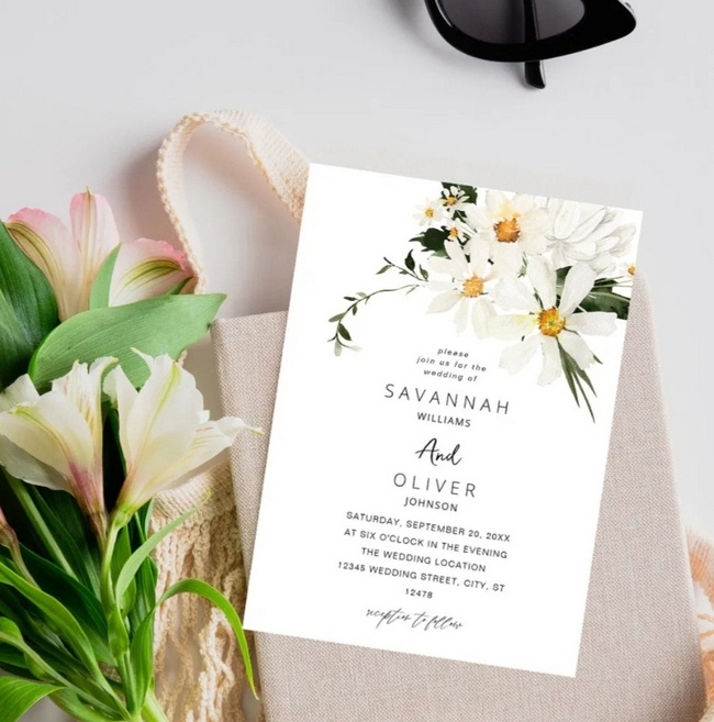 Elegant floral wedding invitation with white daisies and greenery, styled beside fresh flowers and a tote bag.