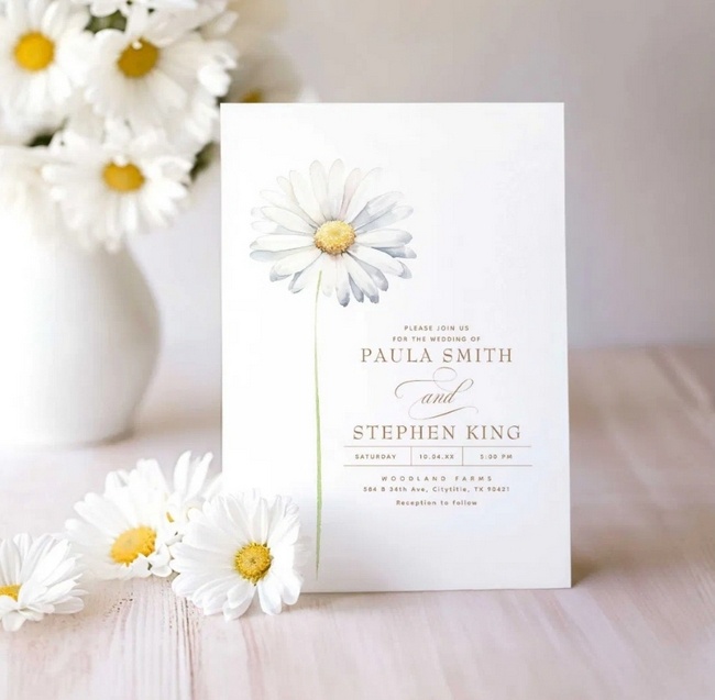 Elegant minimalist wedding invitation with a single white daisy design, styled with fresh daisies in a vase and on the table.