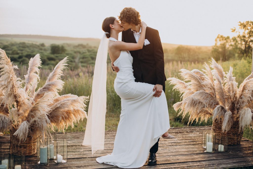 Choose perfect wedding theme romantic outdoor wedding ceremony with bride and groom kissing, pampas grass decorations and sunset boho setting