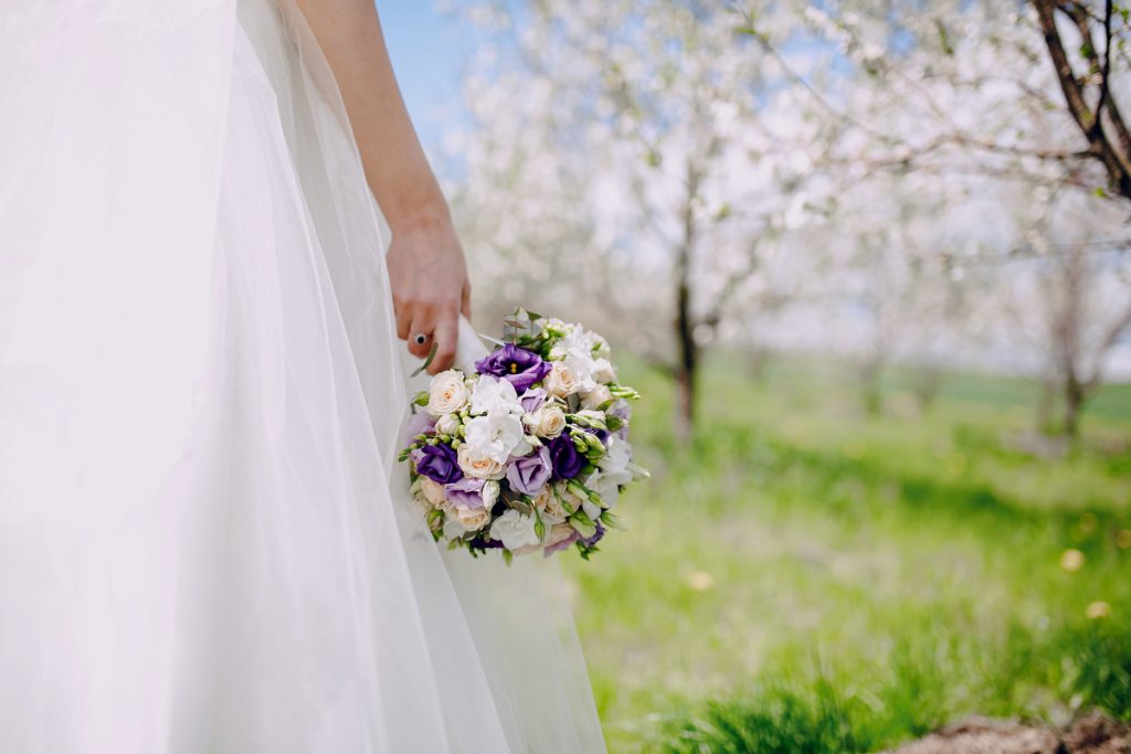 Spring Weddings 2026 bride holding purple and white bouquet in outdoor garden with blooming trees and soft natural light